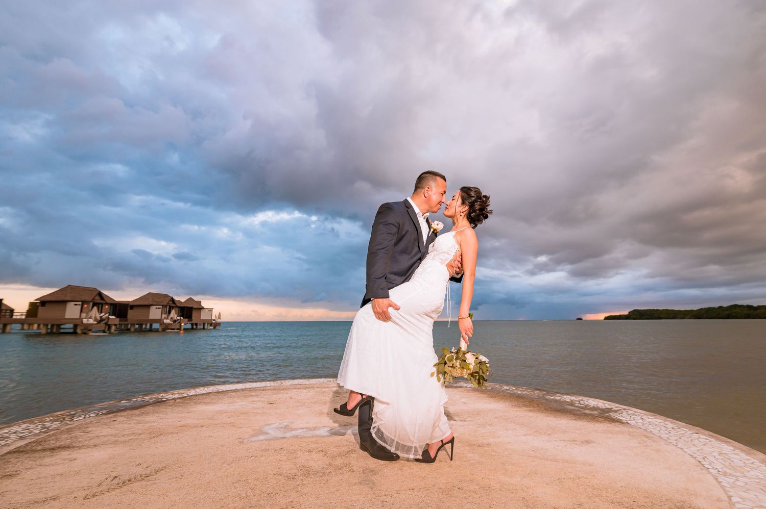 Wedding couple embraces on beach peninsula with overwater villas and dramatic sky in background.