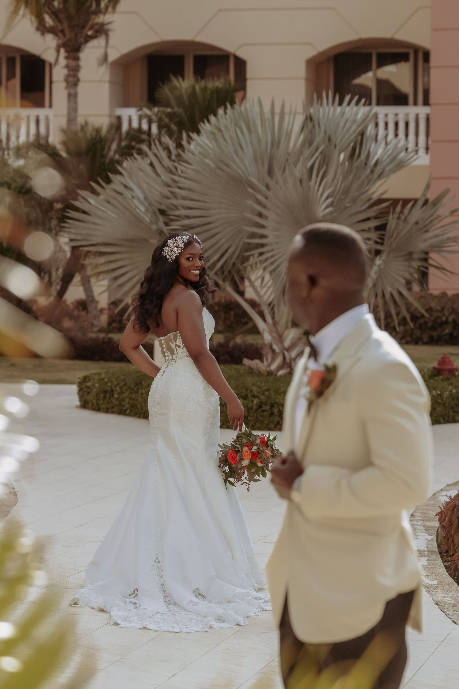 Elegant wedding couple posing outside pink Mediterranean-style building with palm trees.
