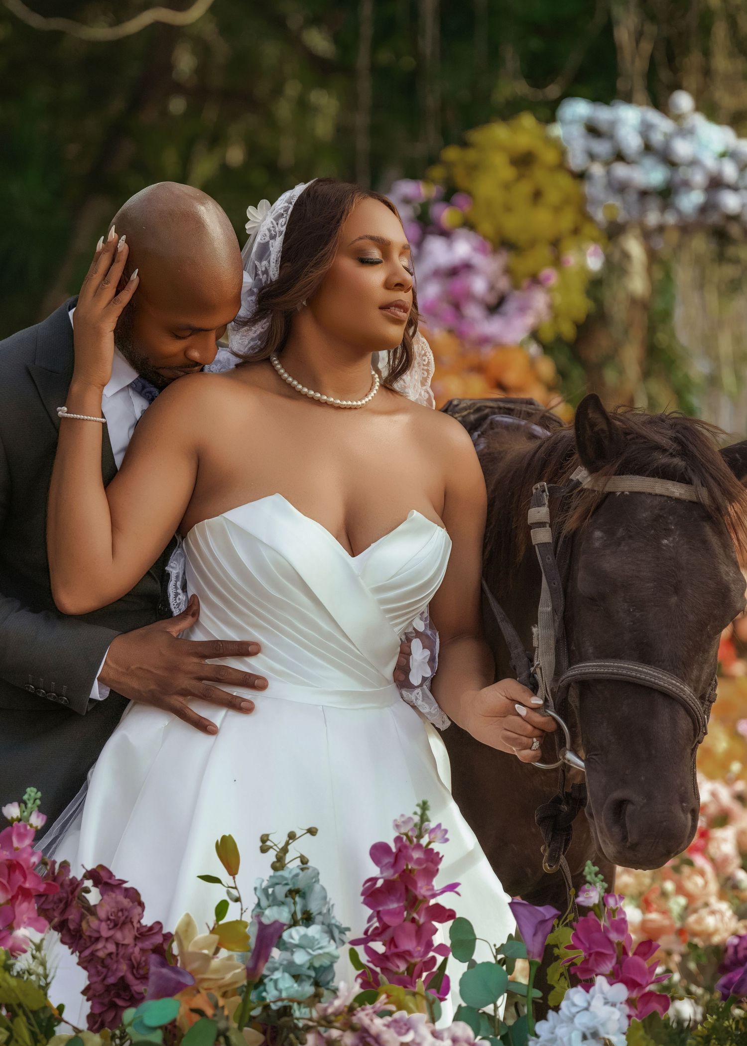 A bride in a white strapless gown poses with a brown horse surrounded by vibrant purple and blue flowers.