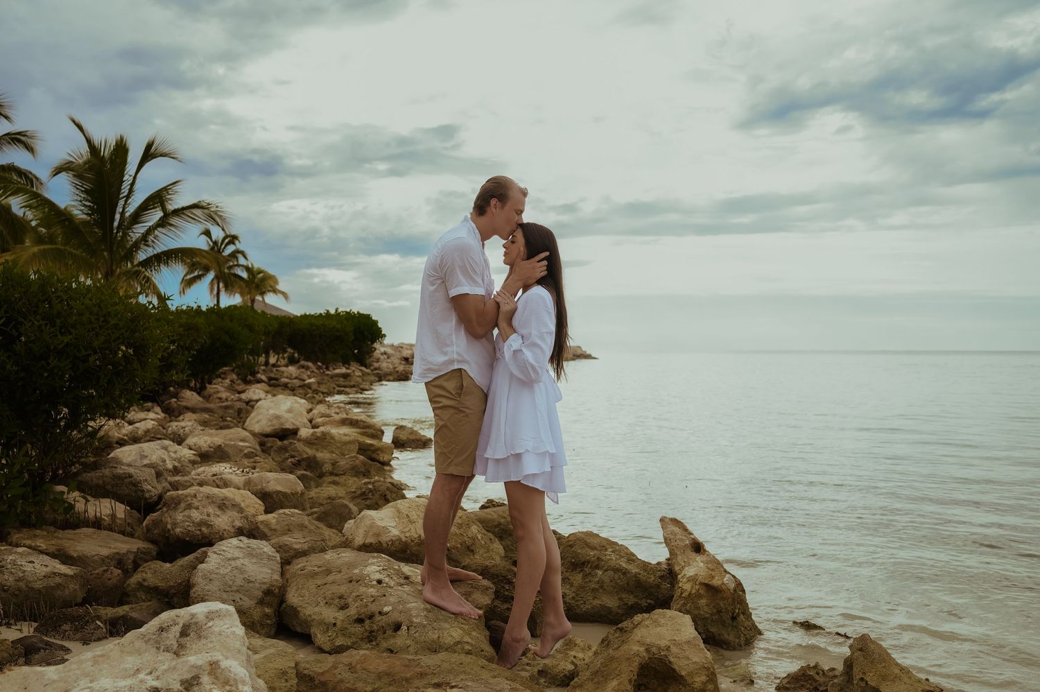 Couple in white and khaki share romantic moments on rocky coastline during sunset.