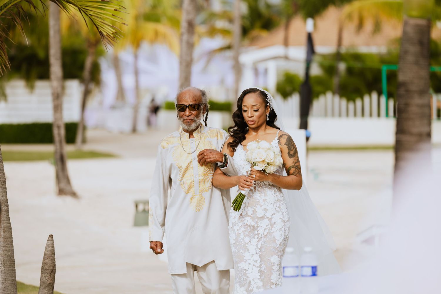 Wedding party members walk along white-railed beachfront path under palm trees.