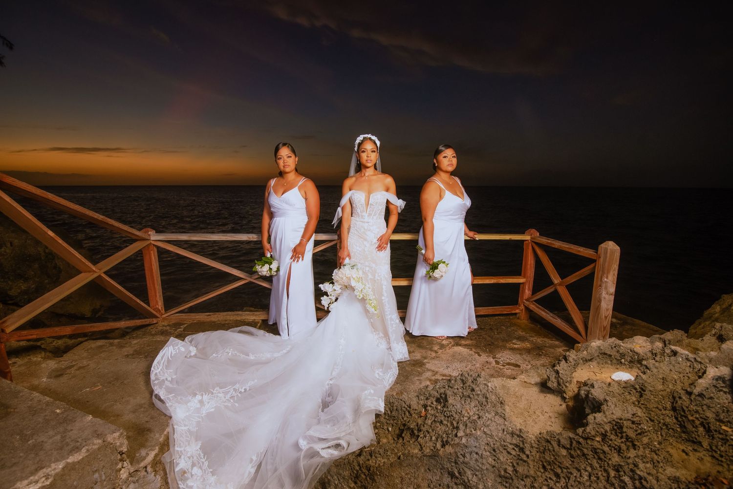 Three bridesmaids in white dresses pose with bride's flowing gown against sunset ocean view.