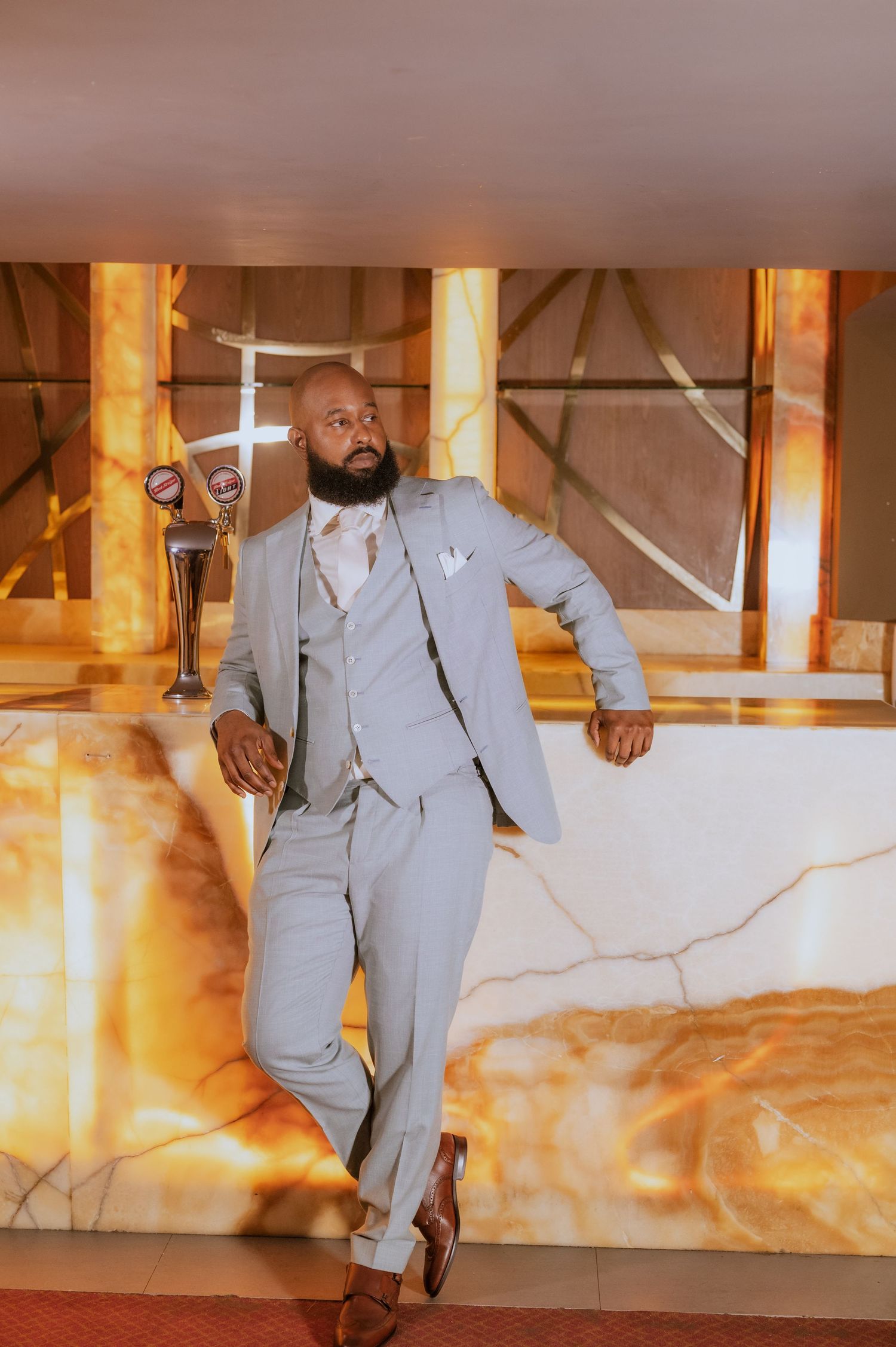Wedding party member in gray suit poses confidently against illuminated marble wall.