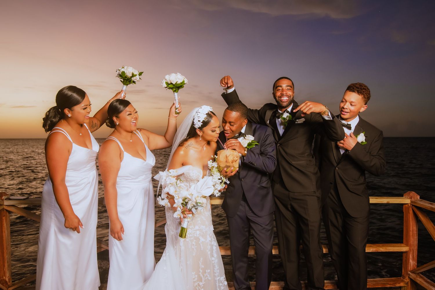 Wedding party celebrates with joyful expressions against dramatic sunset ocean backdrop.