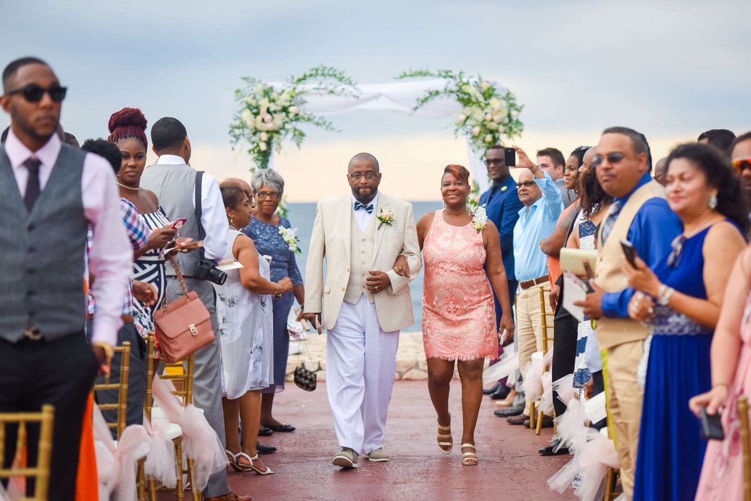 Wedding guests line both sides of an outdoor aisle under a floral arch.