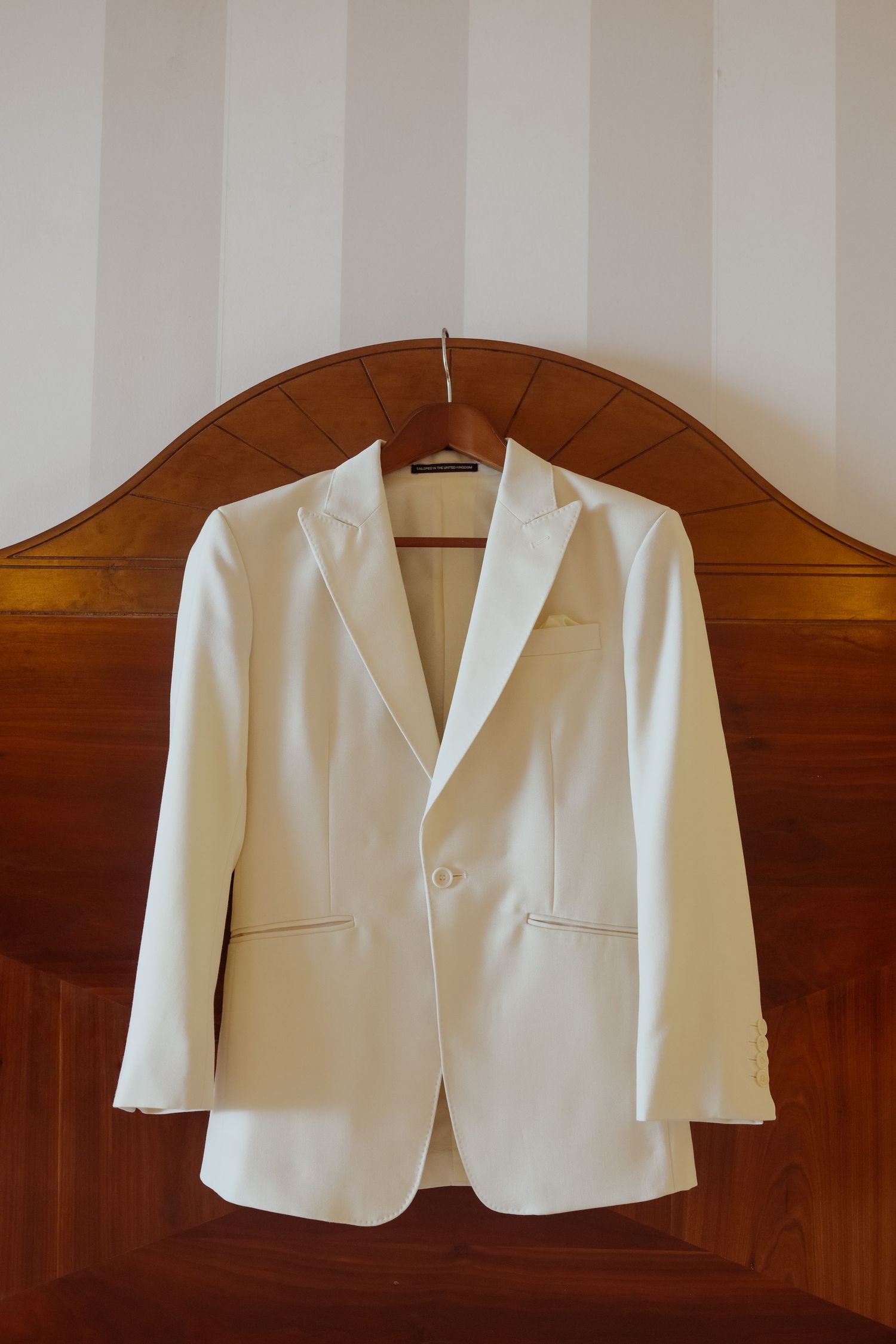 White formal jacket hanging on wooden headboard against striped wallpaper.