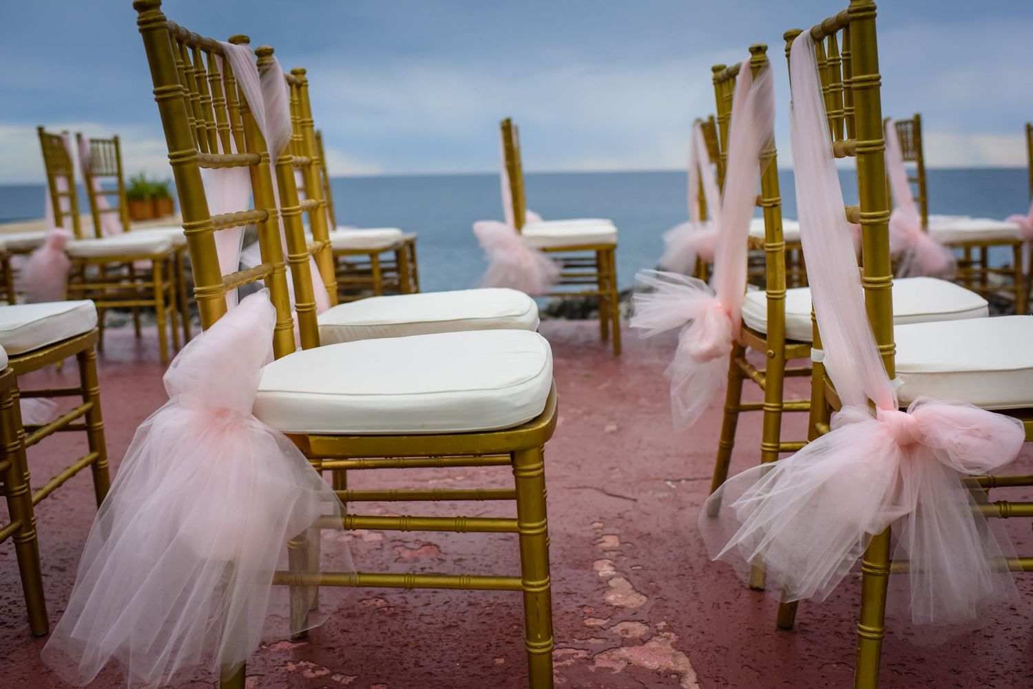 Row of gold chairs decorated with pink tulle bows set up for beach wedding.