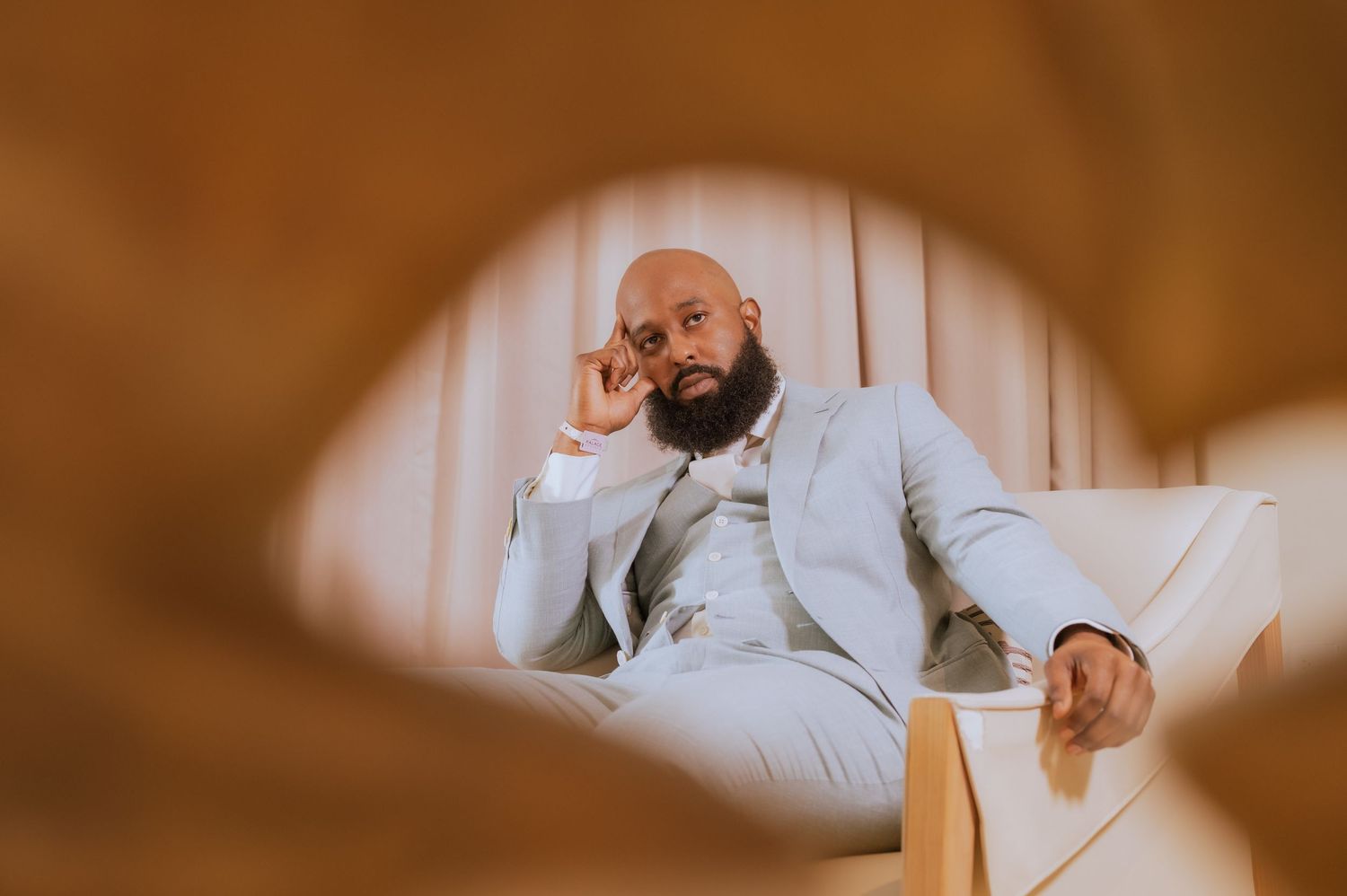 Groom in light gray suit poses thoughtfully on chair through creative frame.