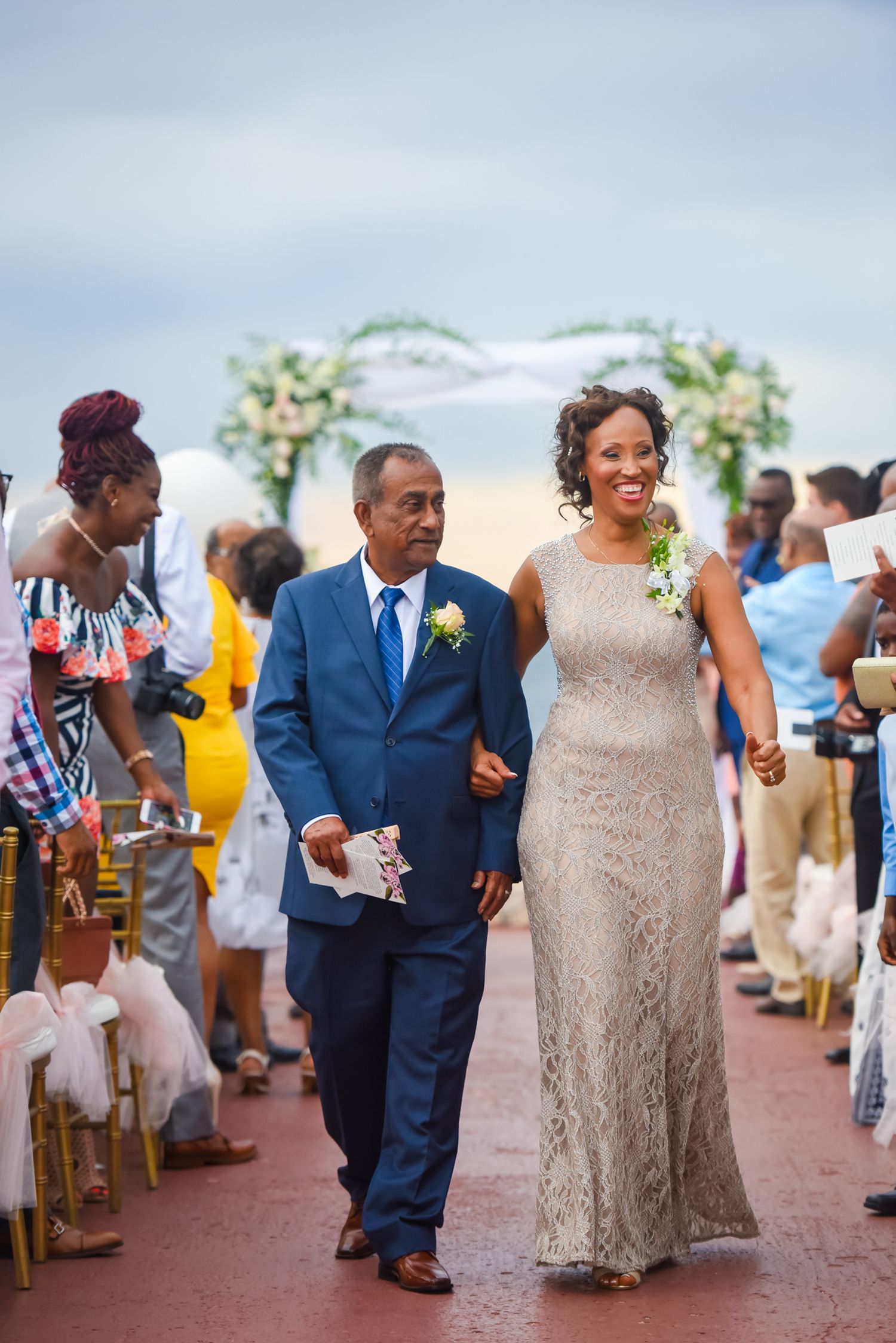 Wedding guests in formal attire walk down an outdoor aisle decorated with floral arrangements.