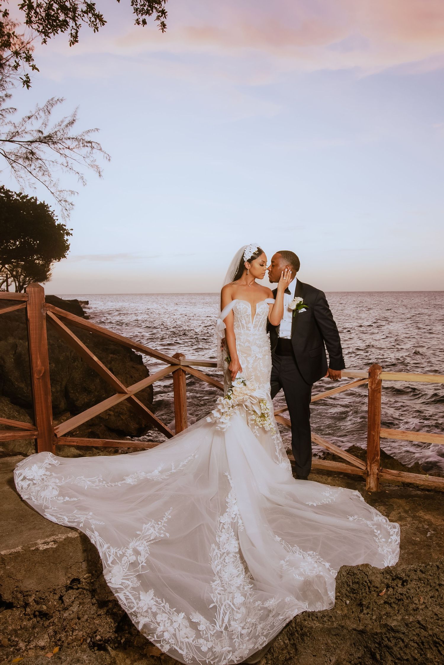 Wedding couple poses along wooden railing at sunset with flowing white dress and ocean view.