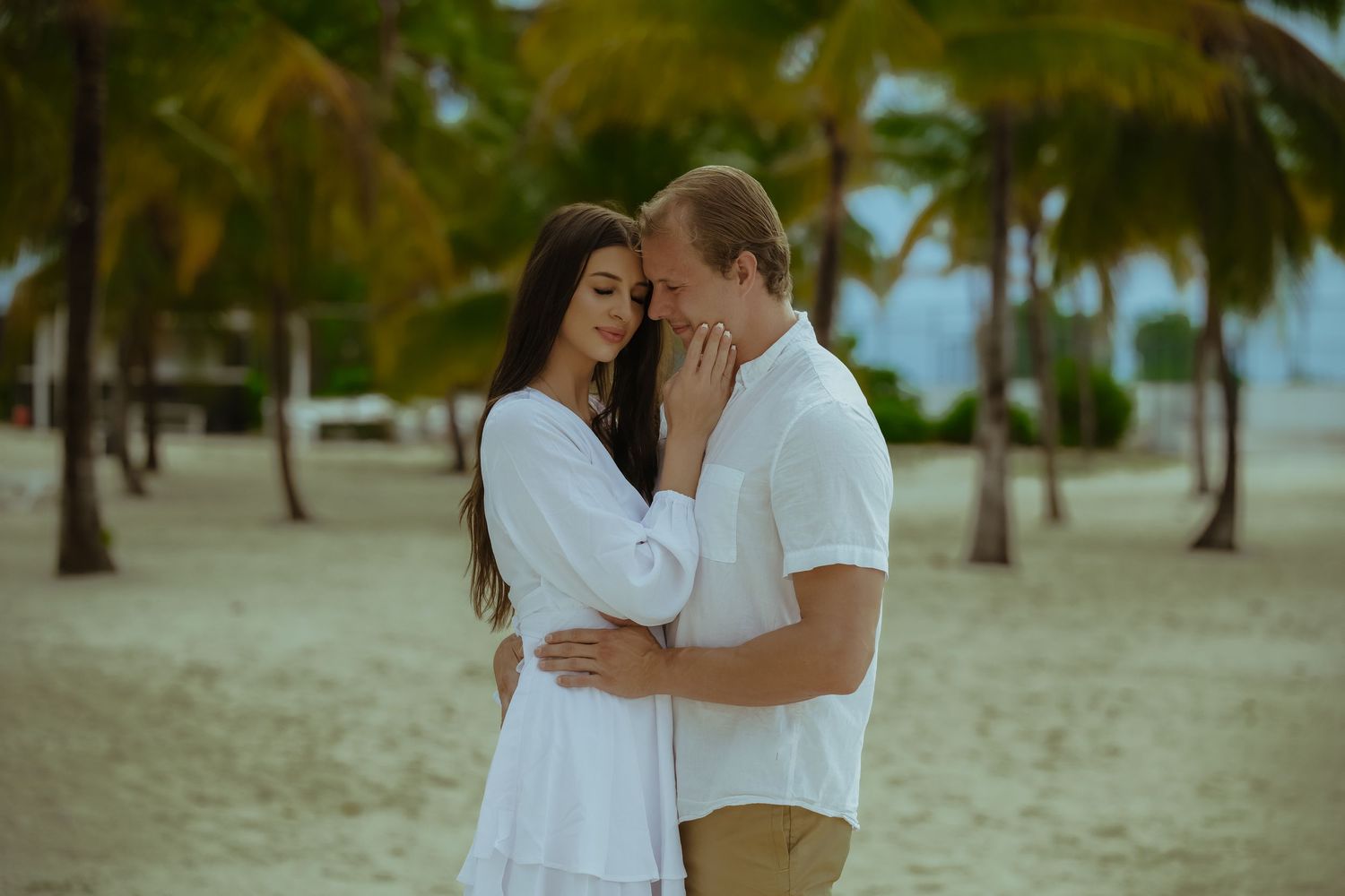 Couple embracing on a sandy beach with palm trees and tropical scenery.