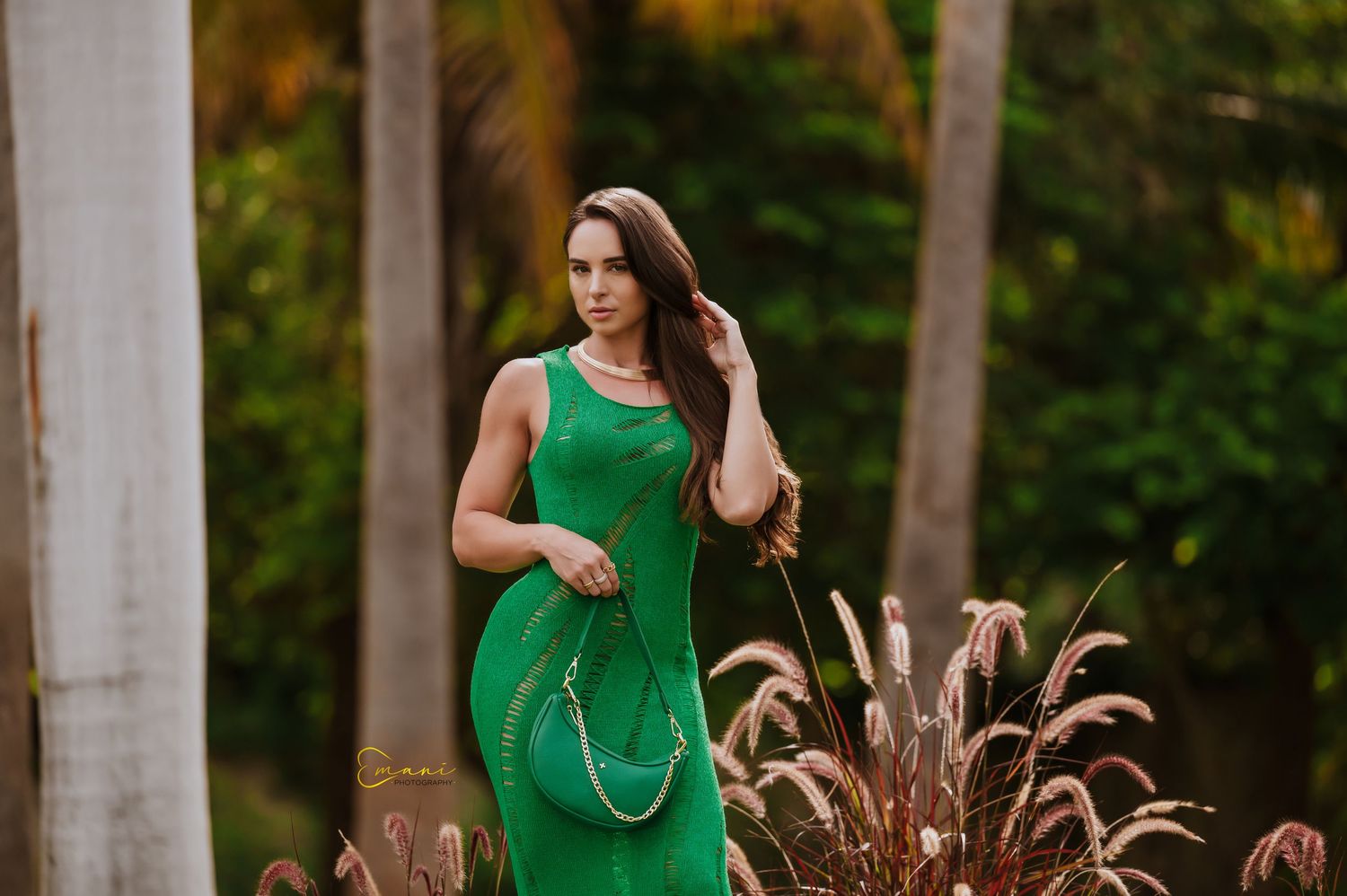 Woman in emerald green dress poses elegantly among tall ornamental grasses at sunset.