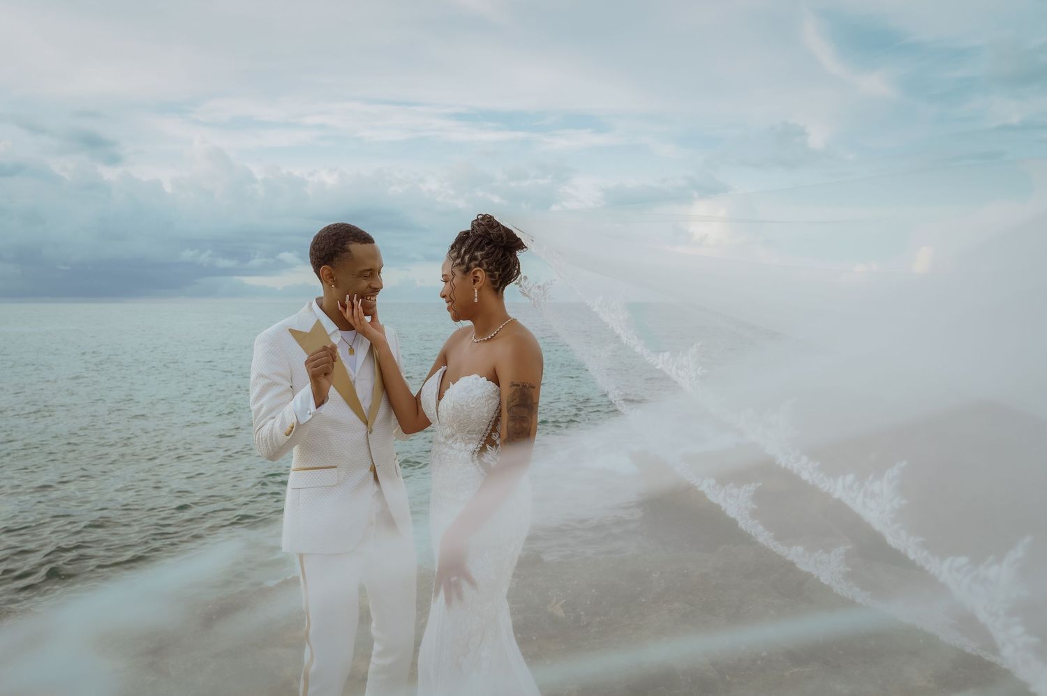 Two people in white wedding attire share a joyful moment together on a cloudy beach as a veil flows in the wind.