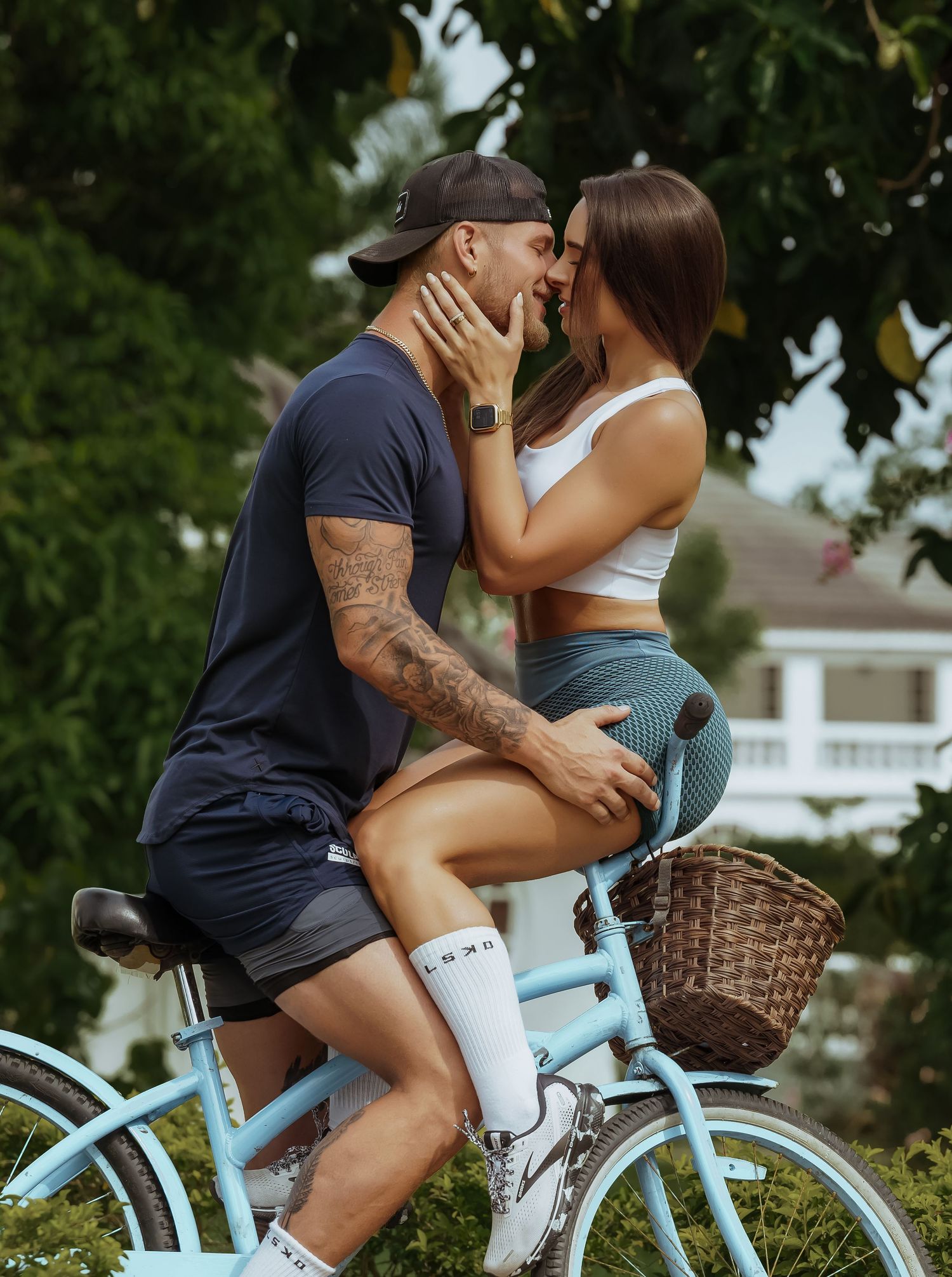 Couple sharing intimate moment on light blue bicycle against garden backdrop.