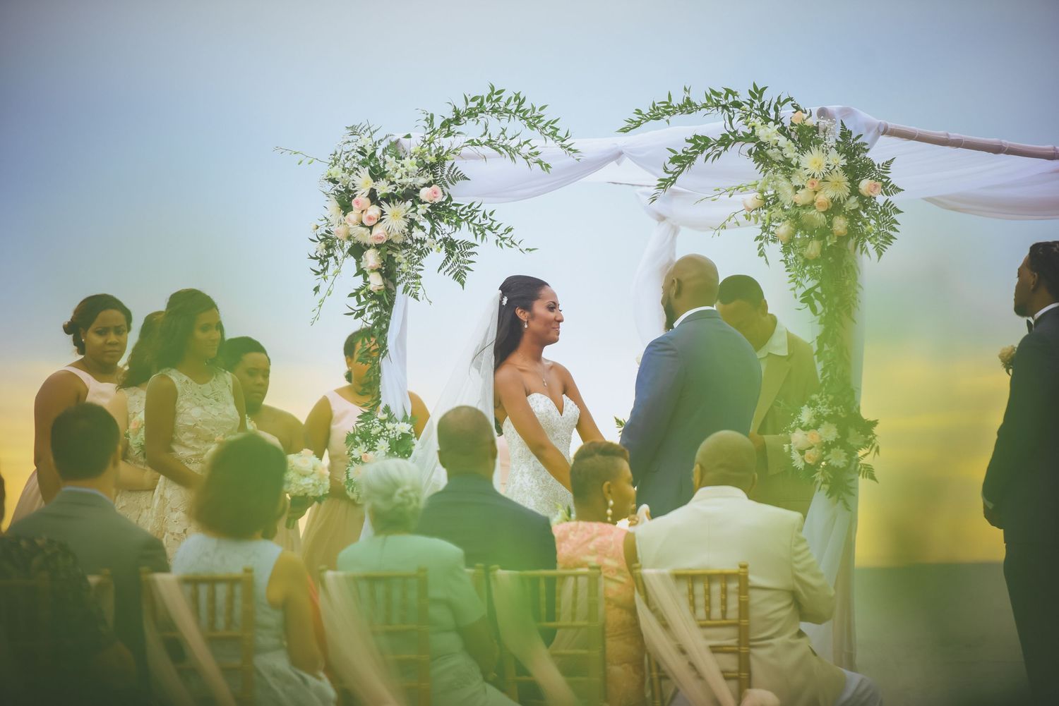 Silhouettes of wedding ceremony participants gather under floral arch during sunset celebration.