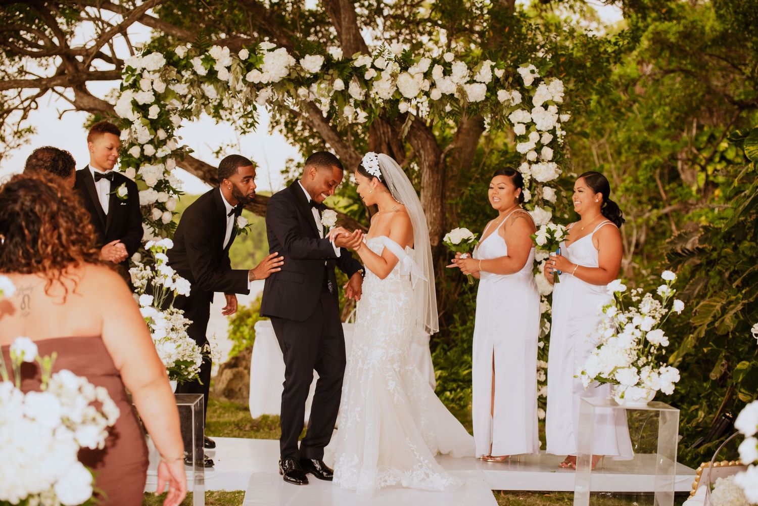 Wedding ceremony scene with white floral arch and guests gathered under trees.