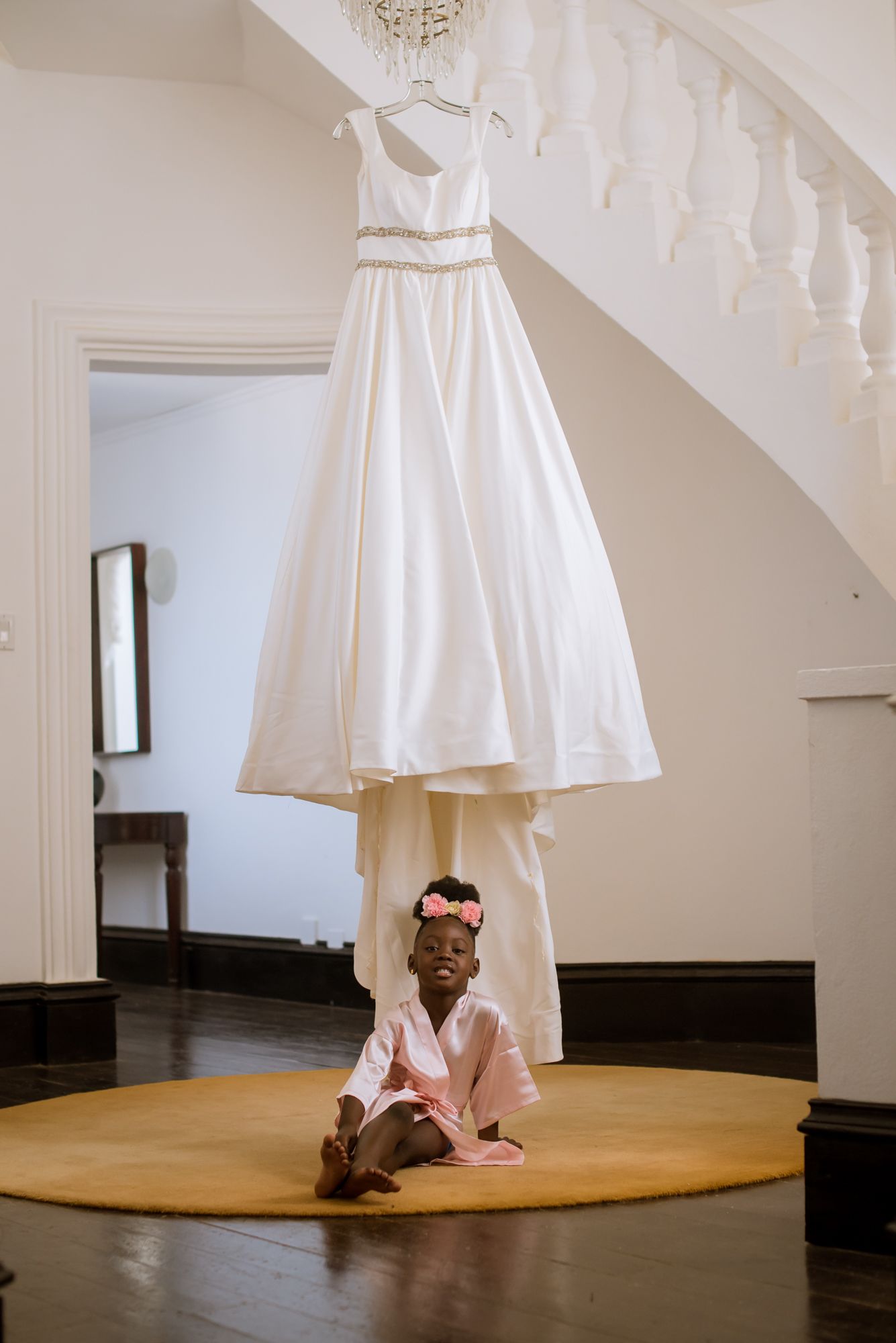 An elegant white wedding dress hangs above a round beige rug near a spiral staircase.