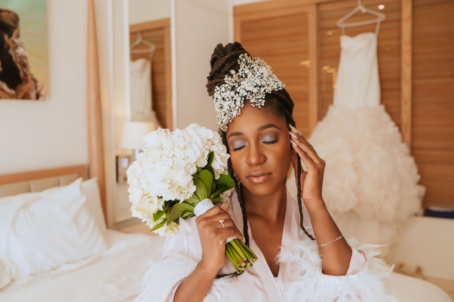 Bride in white robe poses with hydrangea bouquet while wearing crystal headpiece.