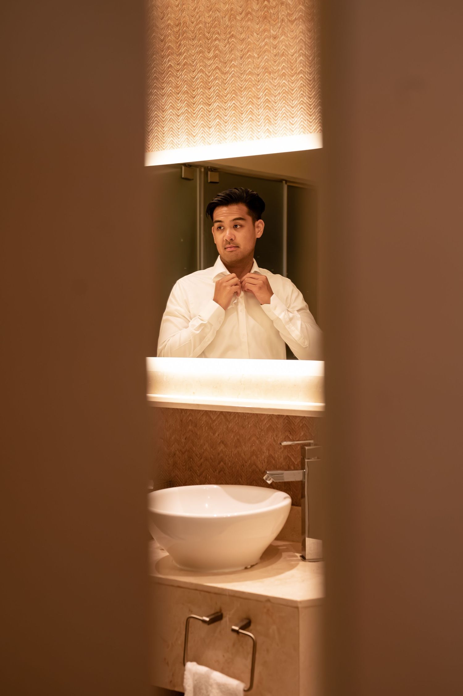 Groom getting ready in bathroom with warm lighting and white sink fixtures.