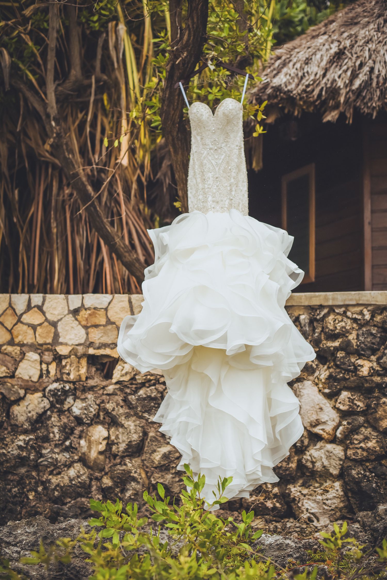 White wedding dress hanging outdoors against tropical foliage and stone wall.