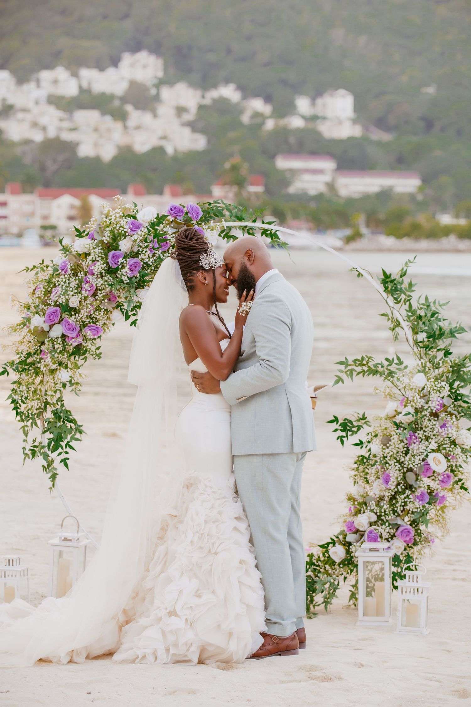 Newlyweds embracing during their beach ceremony with purple floral decorations and white lanterns.