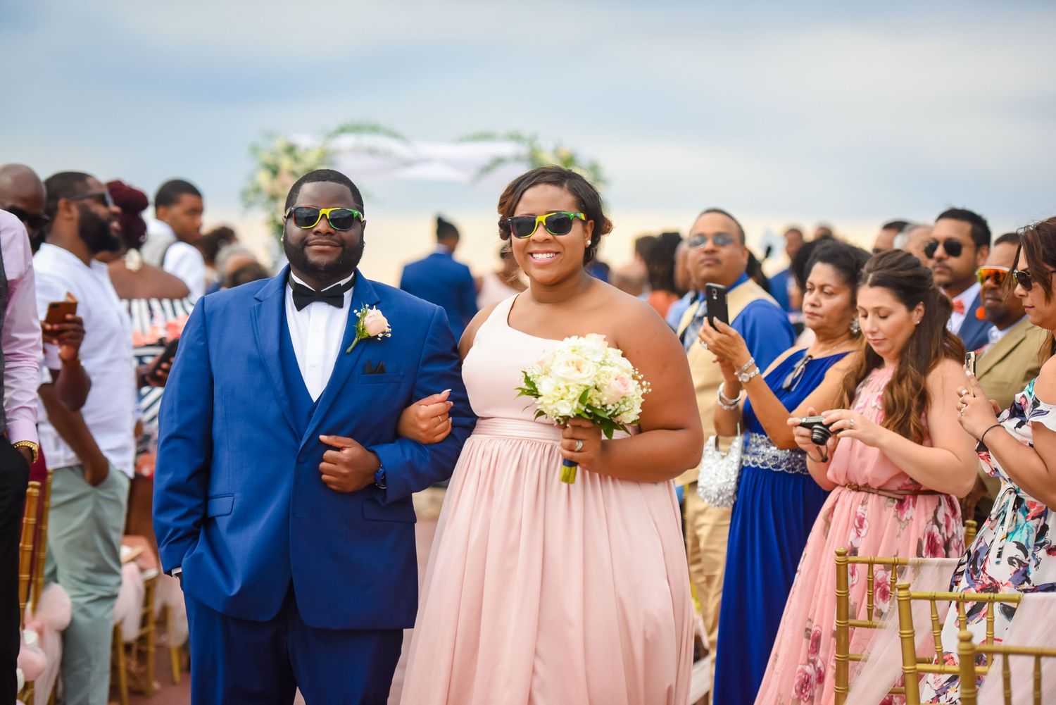 Wedding party in bright blue suits and pink dresses celebrates on a sunny beach.