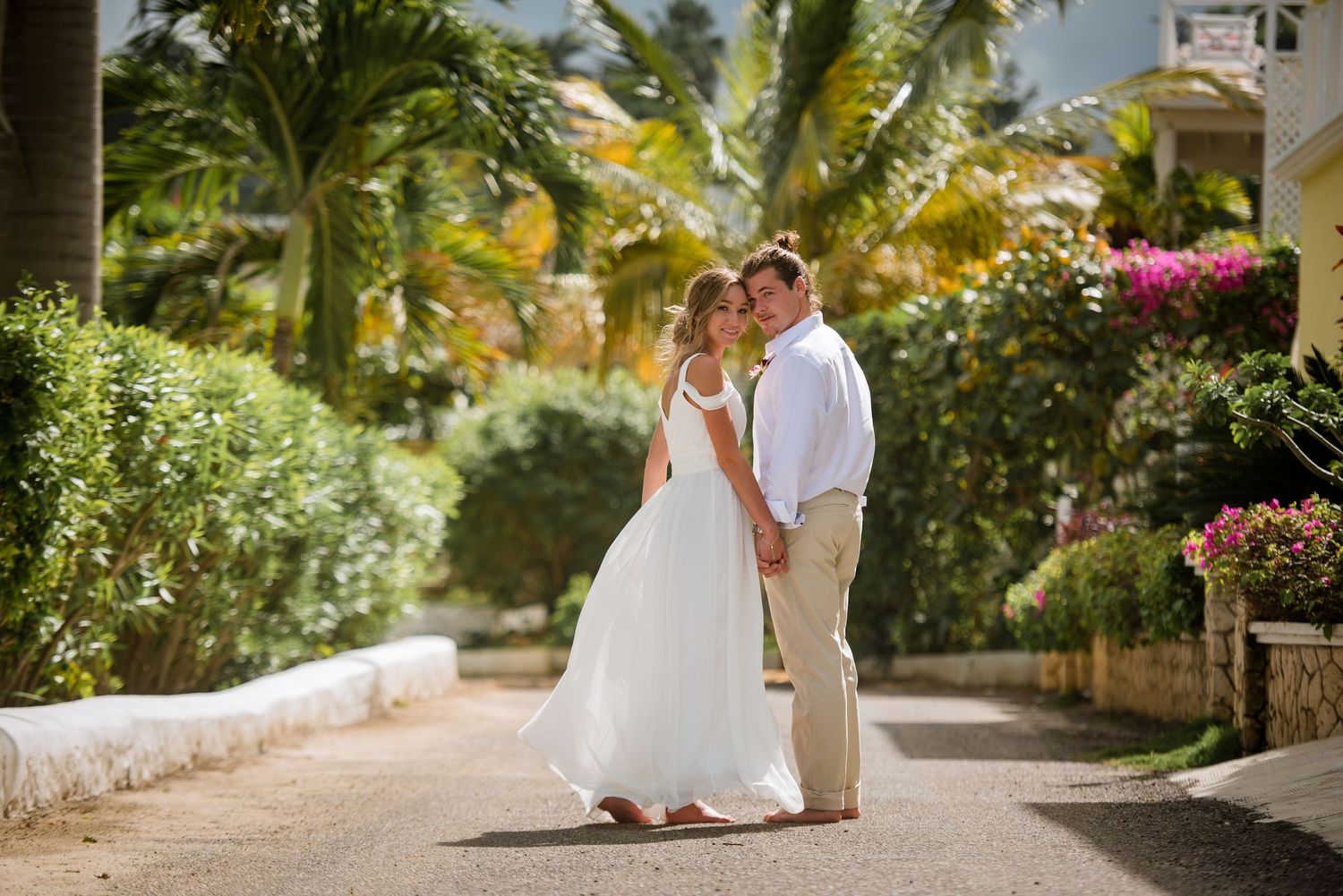 Couple walking along a palm-lined tropical pathway with white walls and flowering plants.