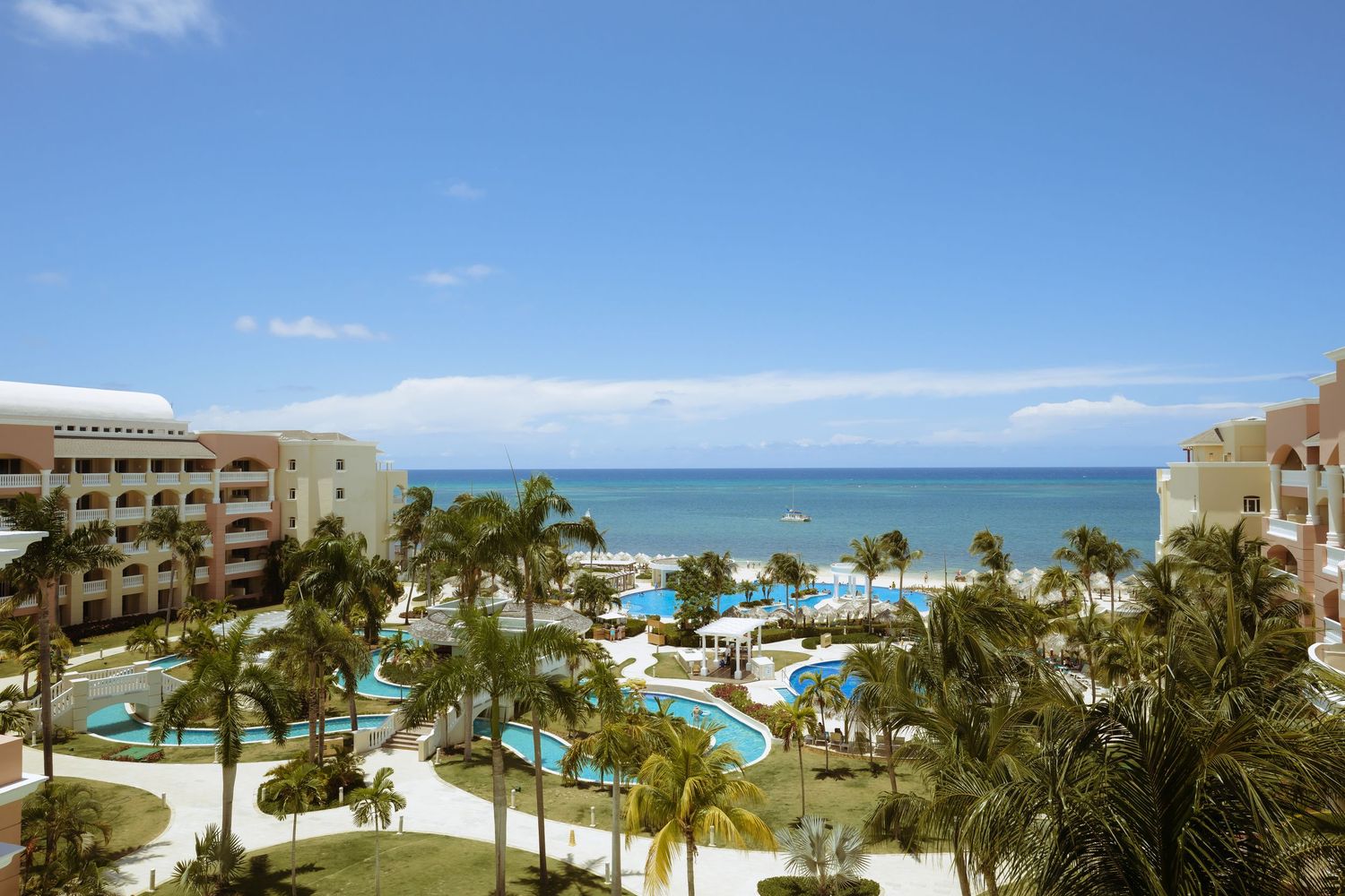 Panoramic view of a beachfront resort with palm trees, swimming pools and turquoise ocean waters under blue skies.