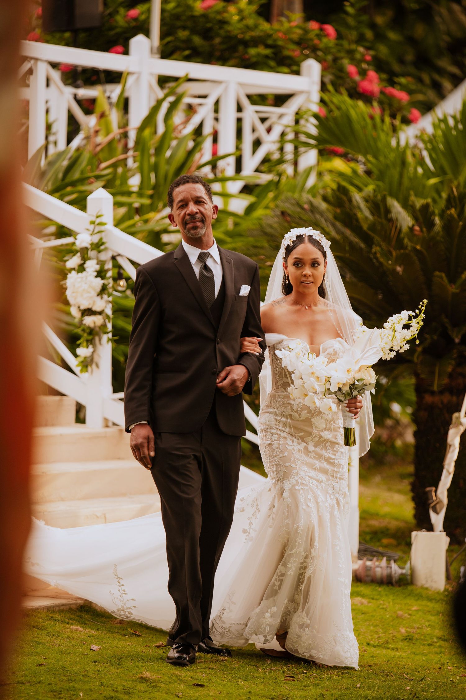 Bride in lace gown walks down garden path with escort during ceremony entrance.
