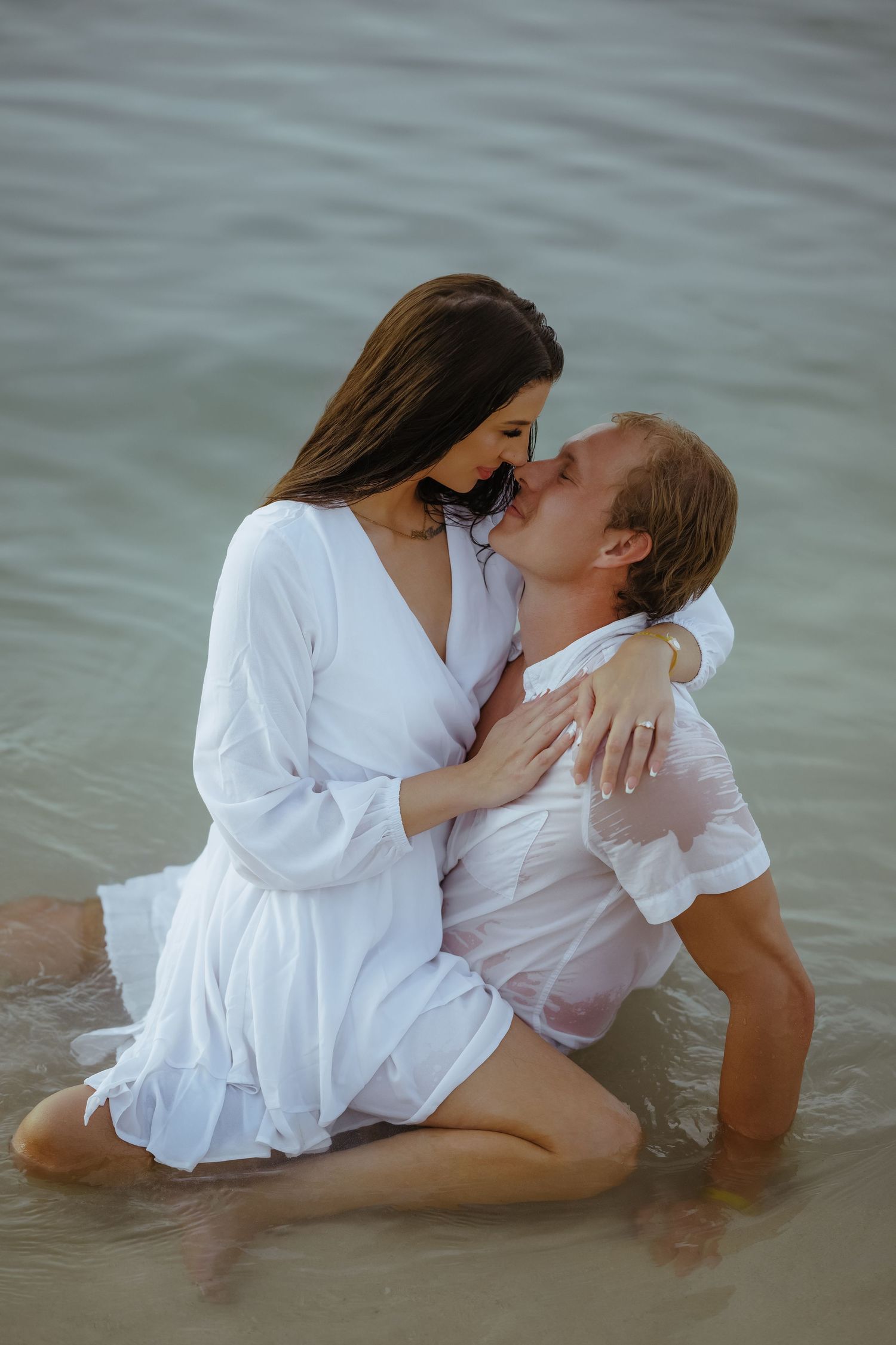 Intimate couple wearing white clothes posing together in ocean waves.