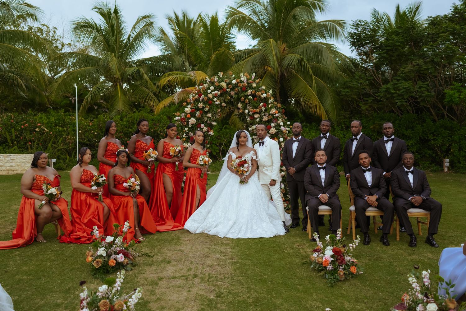 Panoramic wedding party photo with bridesmaids in red dresses against tropical garden backdrop.