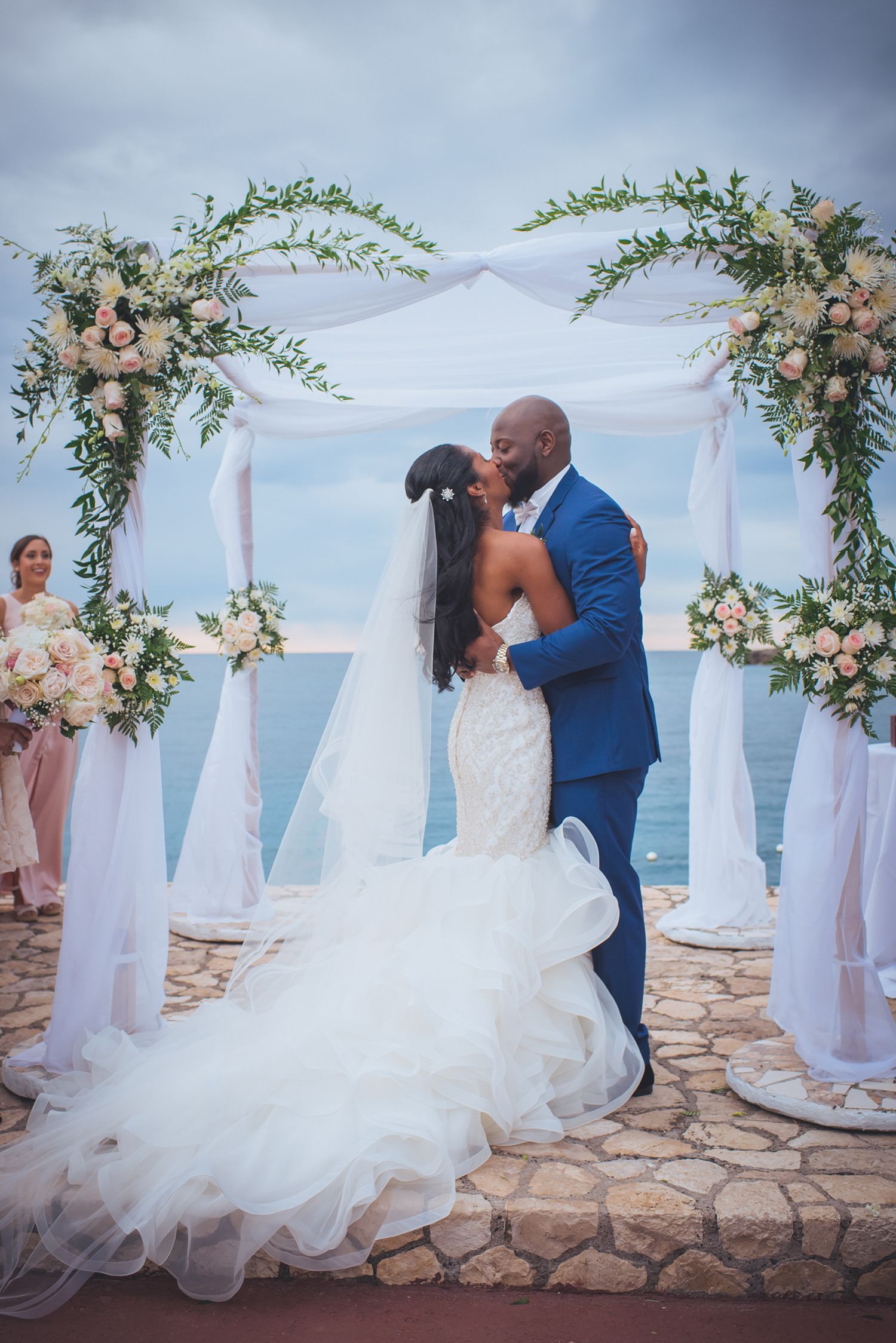 Romantic wedding moment under a floral arch with ocean views and white drapery.