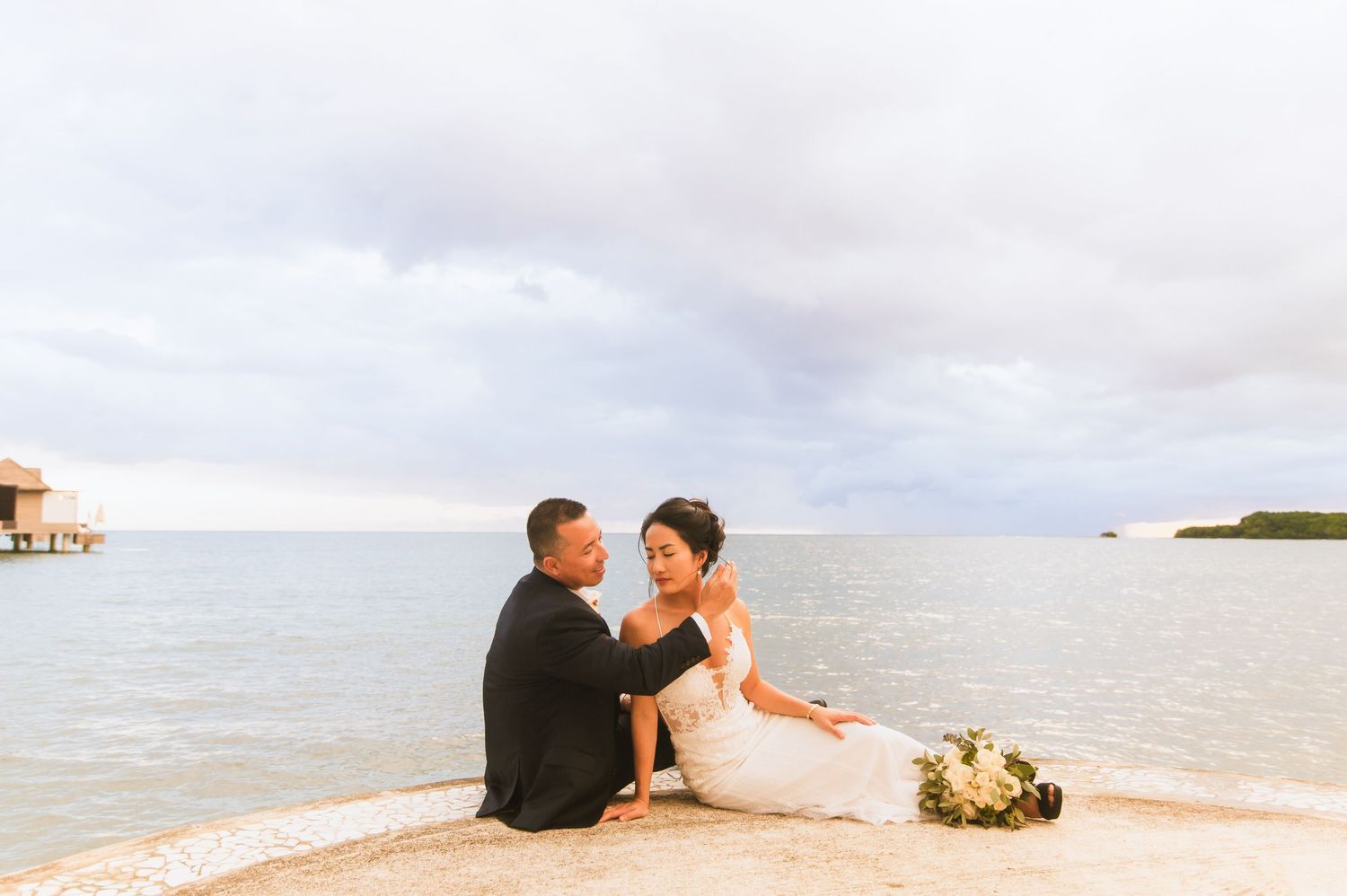 Couple sits together on sandy beach with overwater bungalows visible in background.