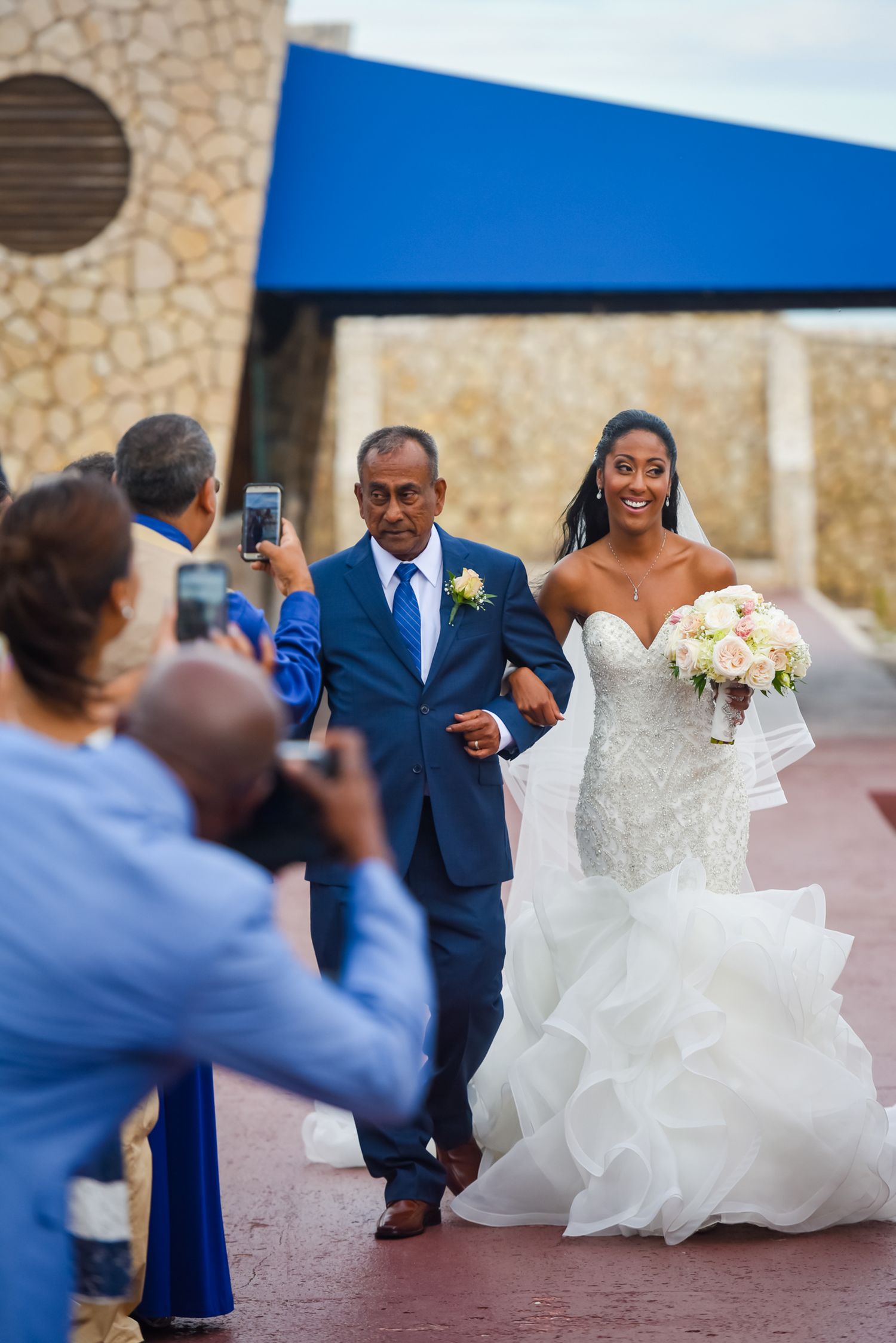 Wedding processional on red walkway with guests taking photos of bride in flowing white gown.