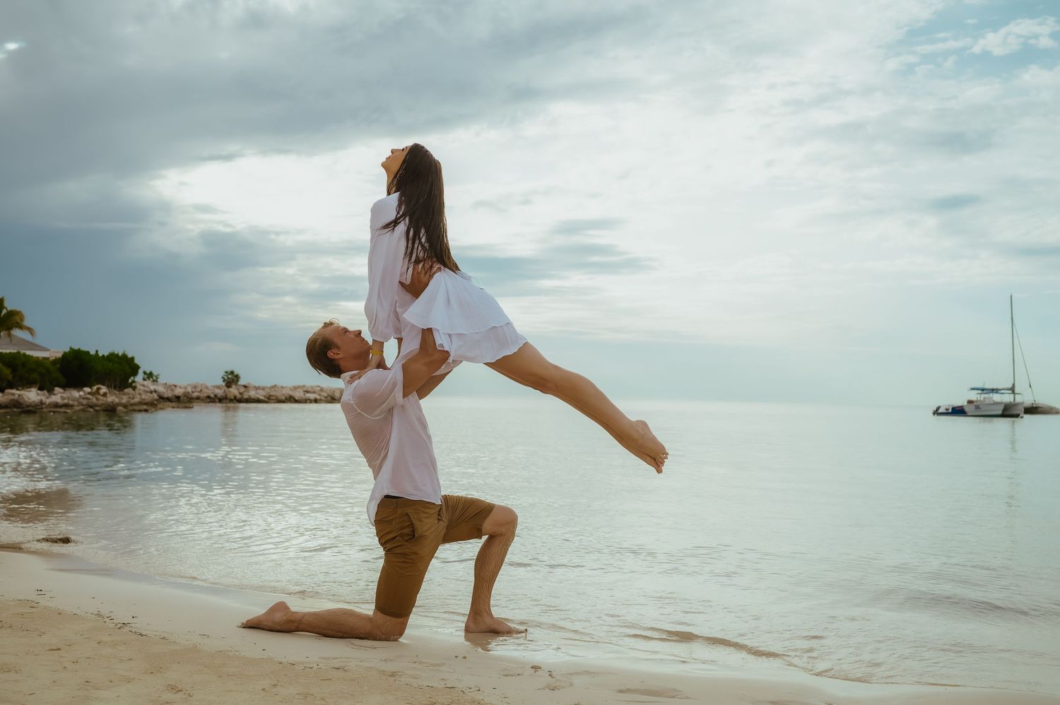 A couple performs a playful lift pose on a tropical beach with boats in the background.