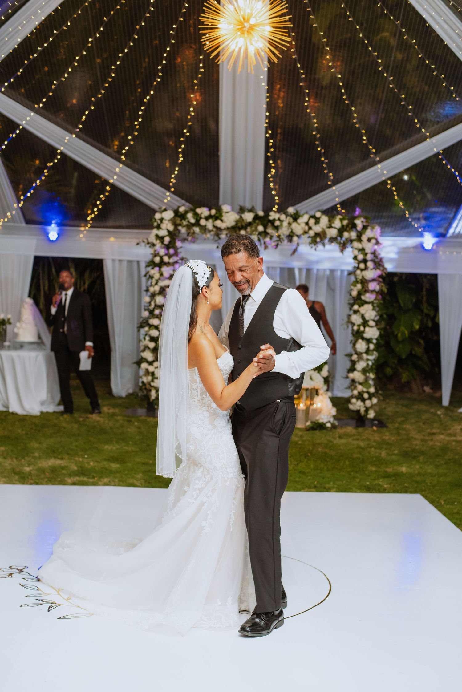 Couple dancing on white dance floor under clear tent with floral arch and twinkling lights.