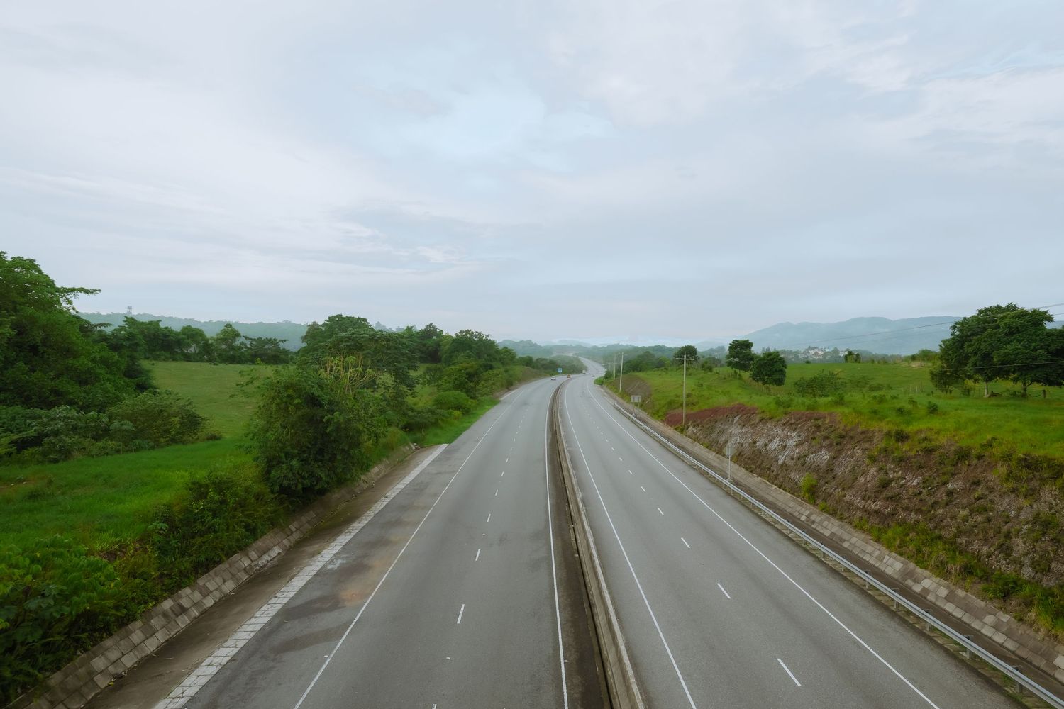 Empty divided highway stretching through green rolling hills under cloudy sky.