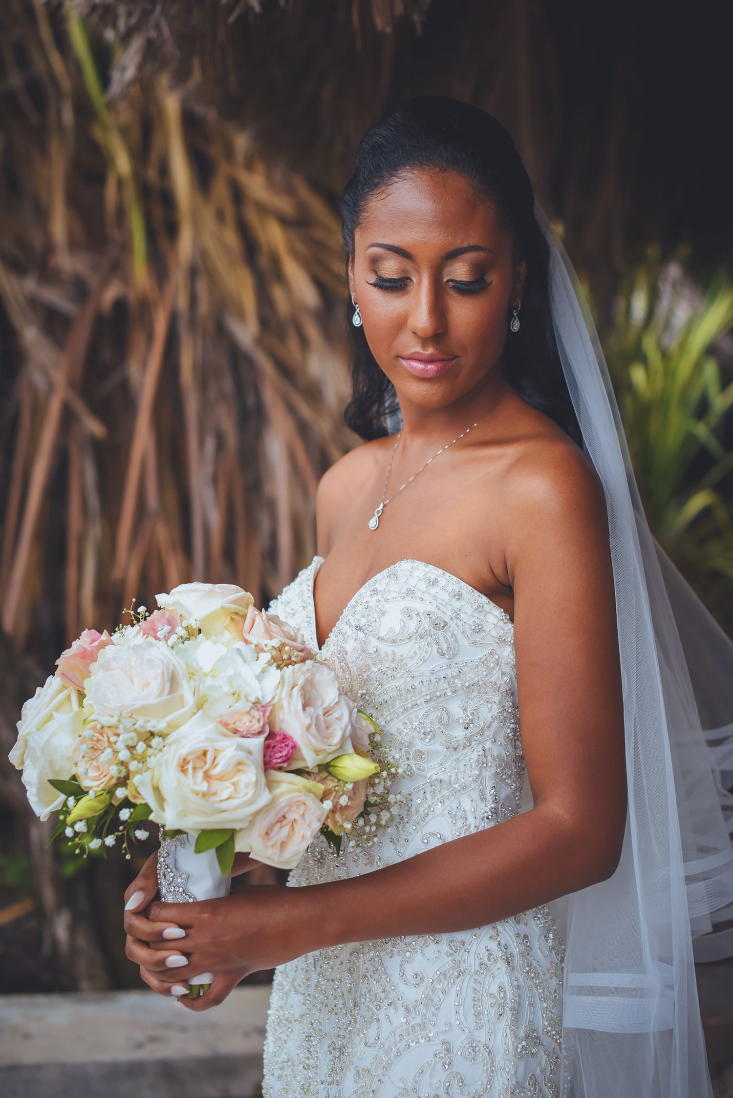 Bride holding an elegant bouquet of white and blush roses against tropical foliage.