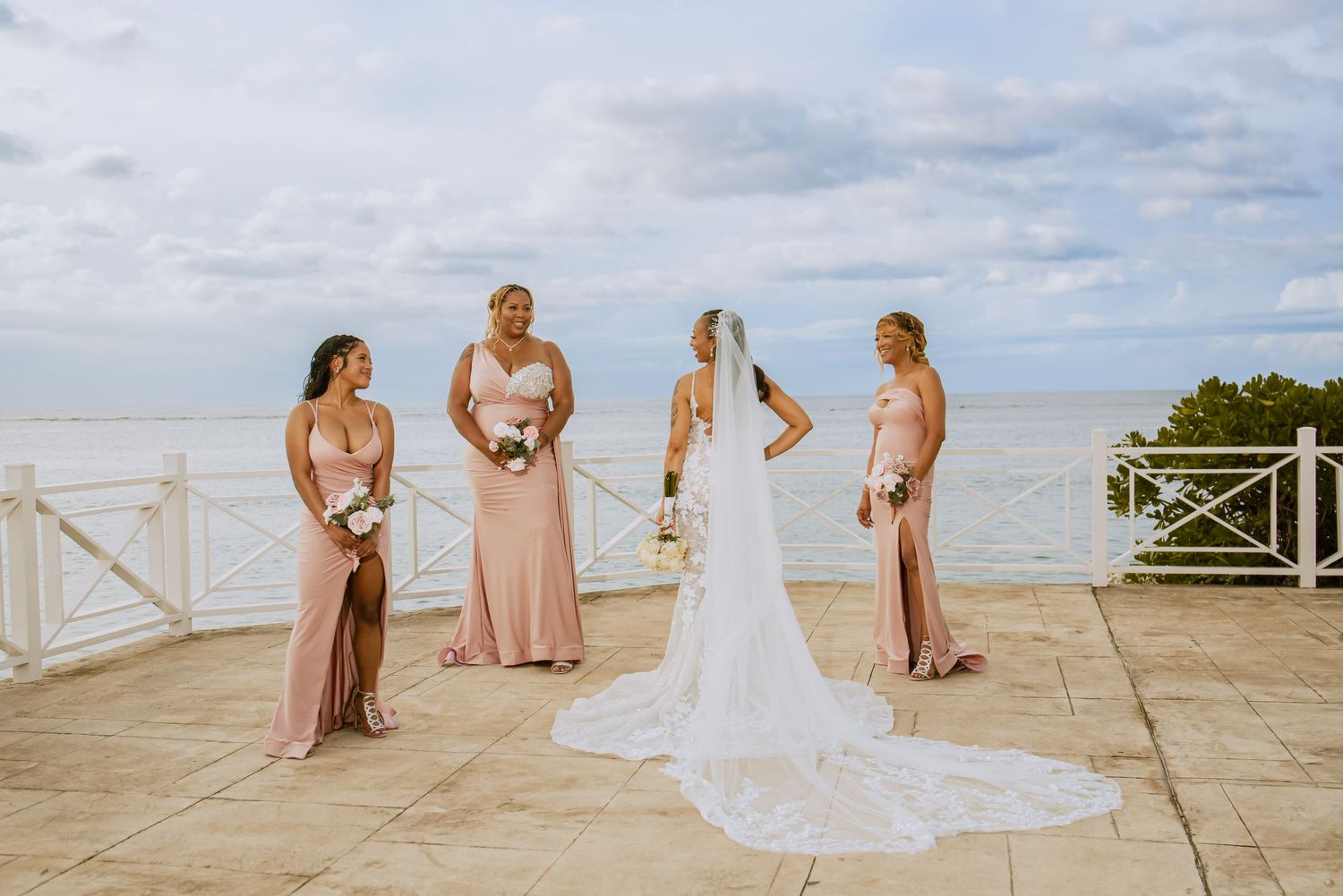 Bride in white gown with long train poses with bridesmaids in pink dresses by the sea.