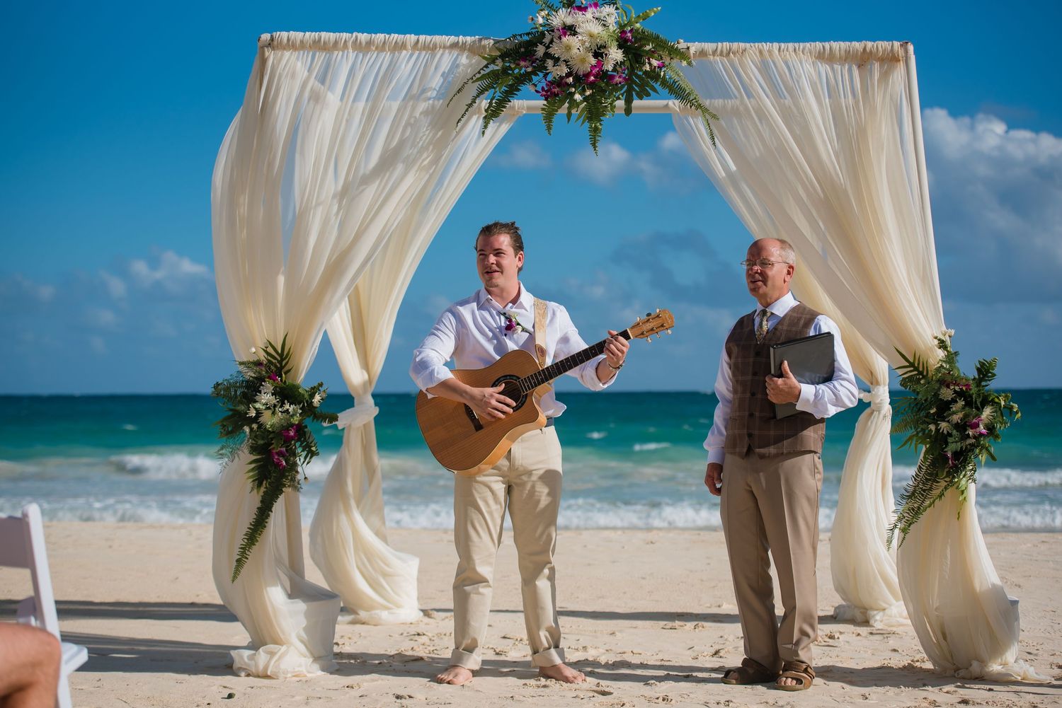 Musicians perform at a beach wedding ceremony under white fabric draped arch.