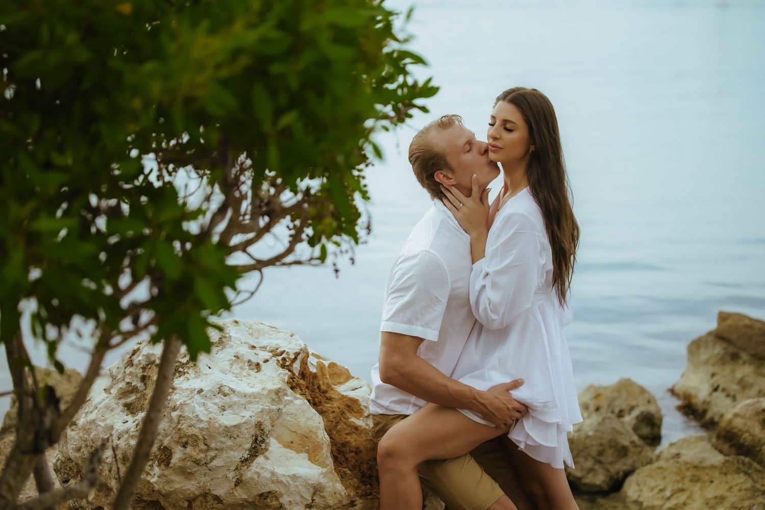 A romantic moment captured between a couple in white outfits on rocky coastal terrain.
