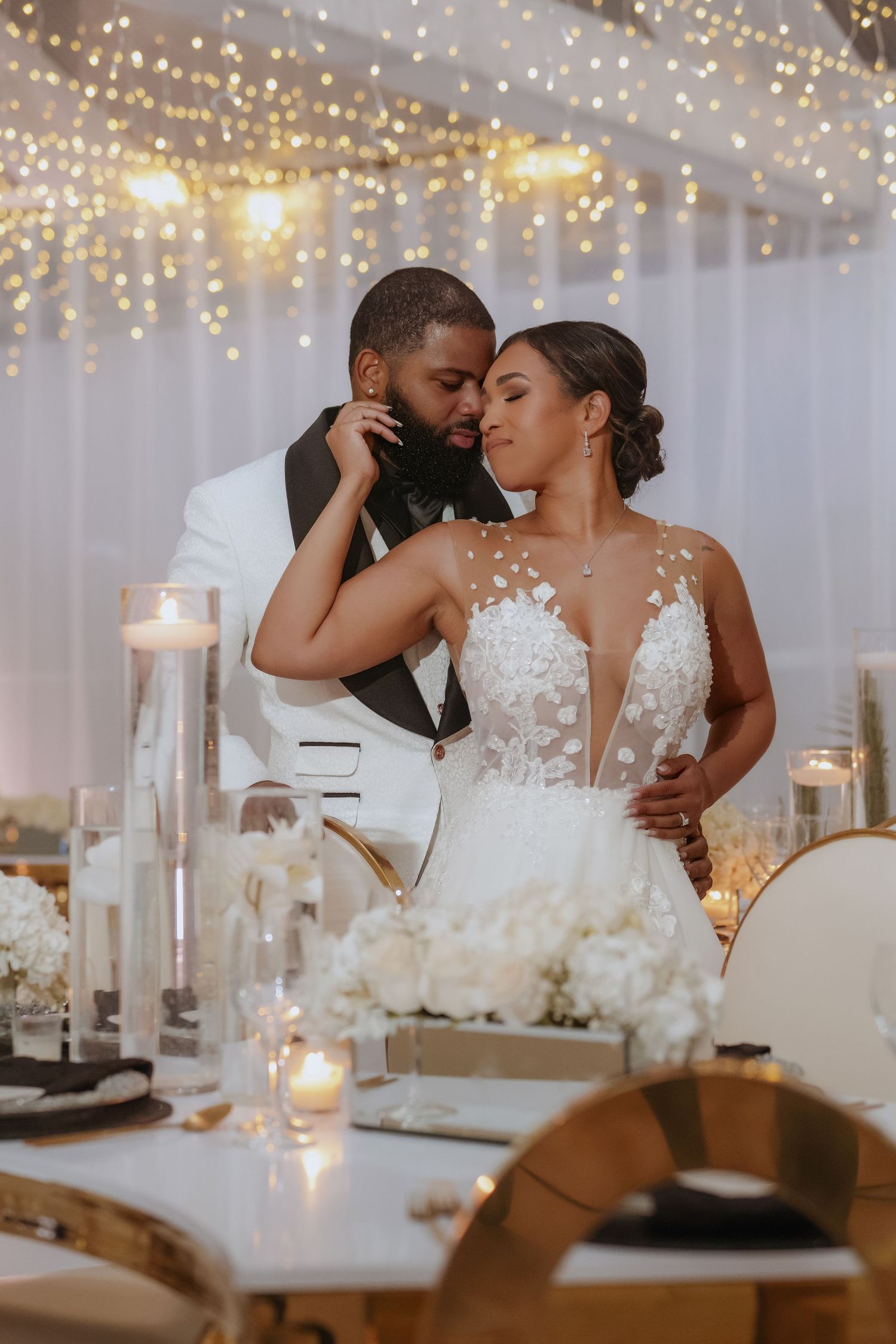 Romantic wedding reception scene with twinkling lights and white decor as newlyweds share intimate moments at their table.