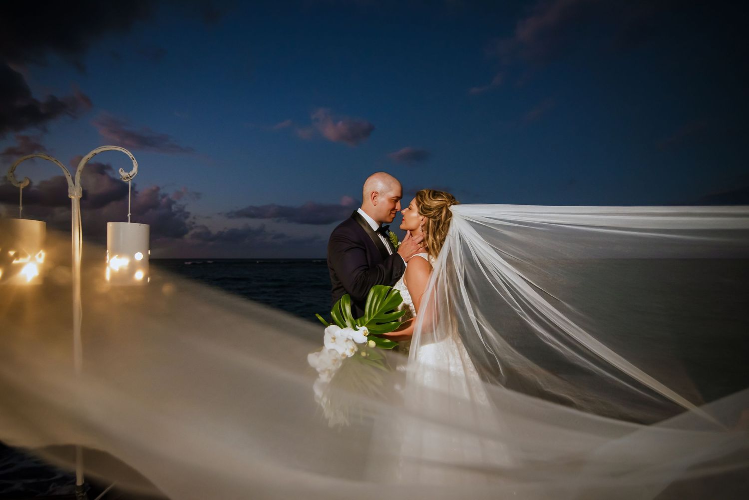 A dramatic wedding photo at sunset with a flowing veil creating an ethereal silhouette against the darkening sky.