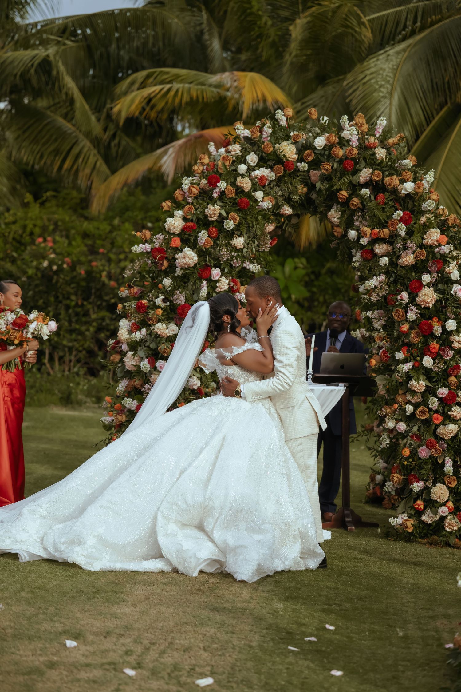 Wedding couple posing on grass with floral arch of red and white roses in tropical garden setting.