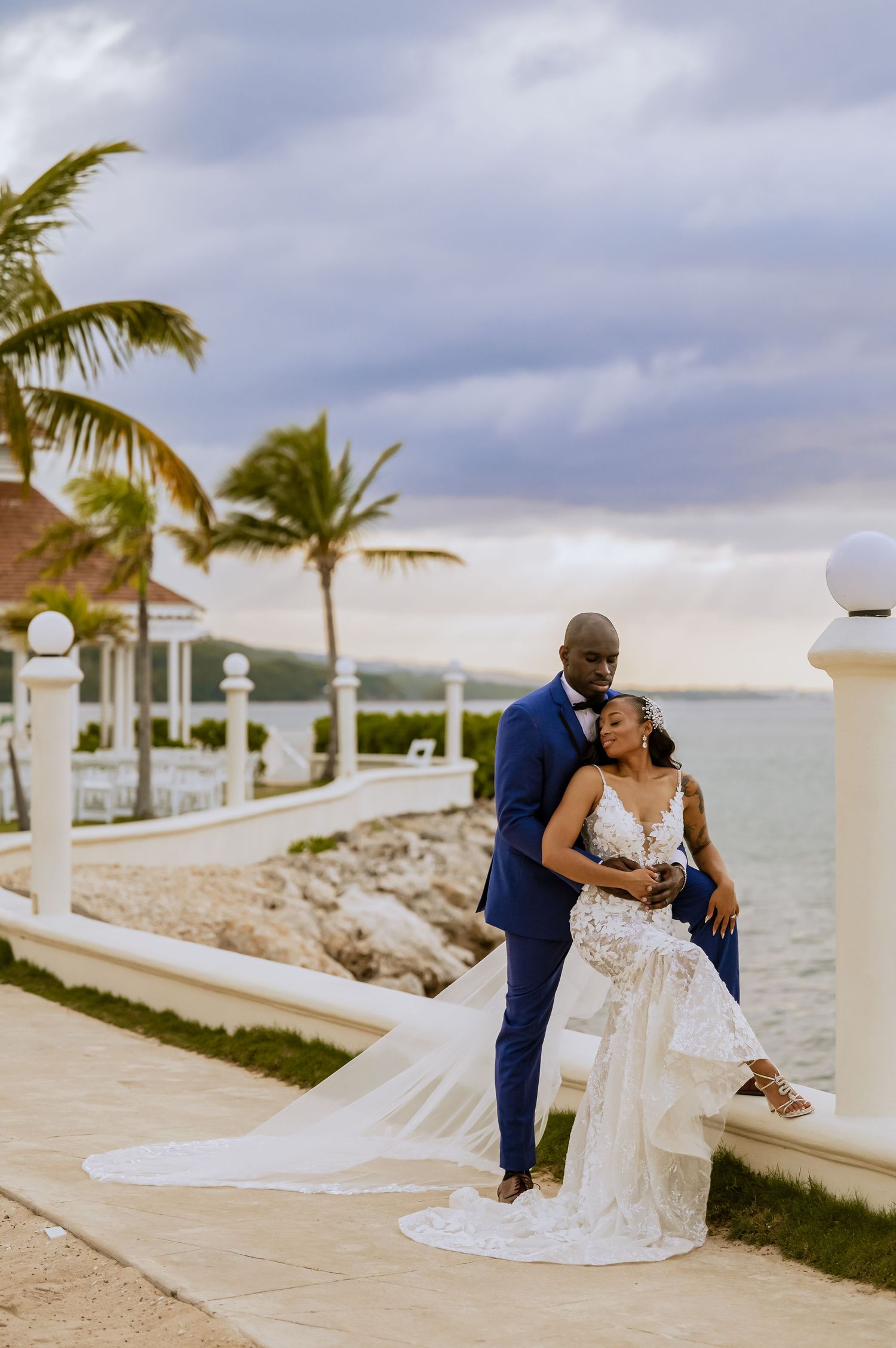 A wedding couple shares an intimate moment on a coastal walkway surrounded by palm trees and ocean views.