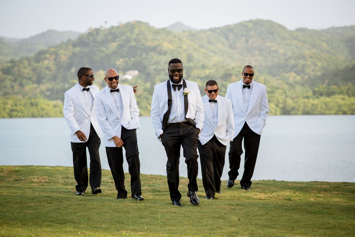 Groomsmen in black suits and white shirts pose together on a grassy hill with mountains in the background.