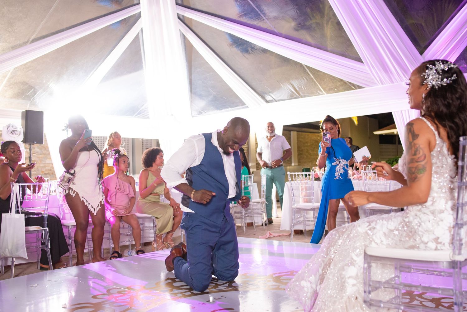 Wedding guests gather on a white dance floor under draped fabric and purple lighting.