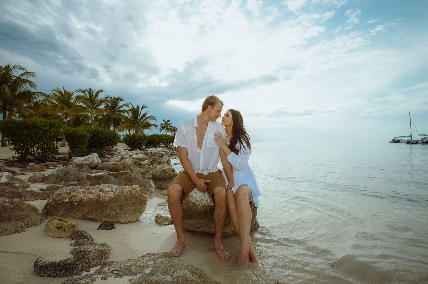 A couple sits together on coastal rocks with a beautiful tropical ocean landscape behind them.