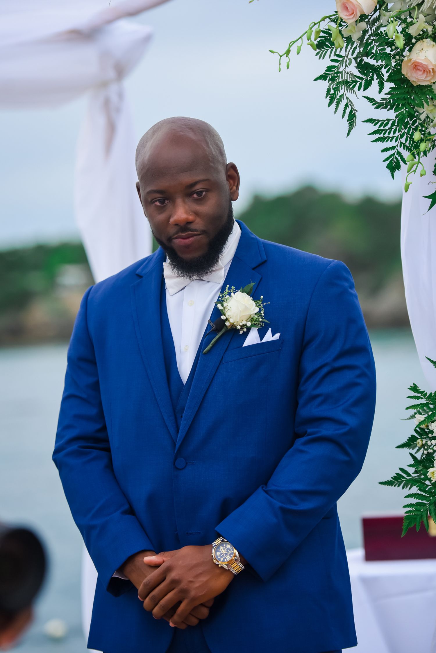 Groom in royal blue three-piece suit stands under floral arch at outdoor ceremony.