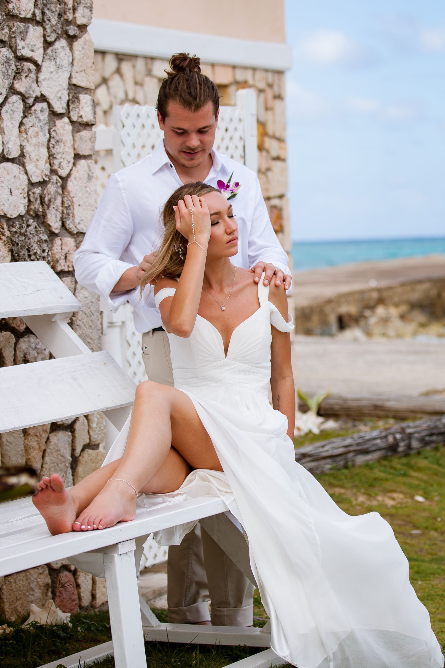Wedding couple sharing intimate moments on white benches against a stone wall with ocean views.